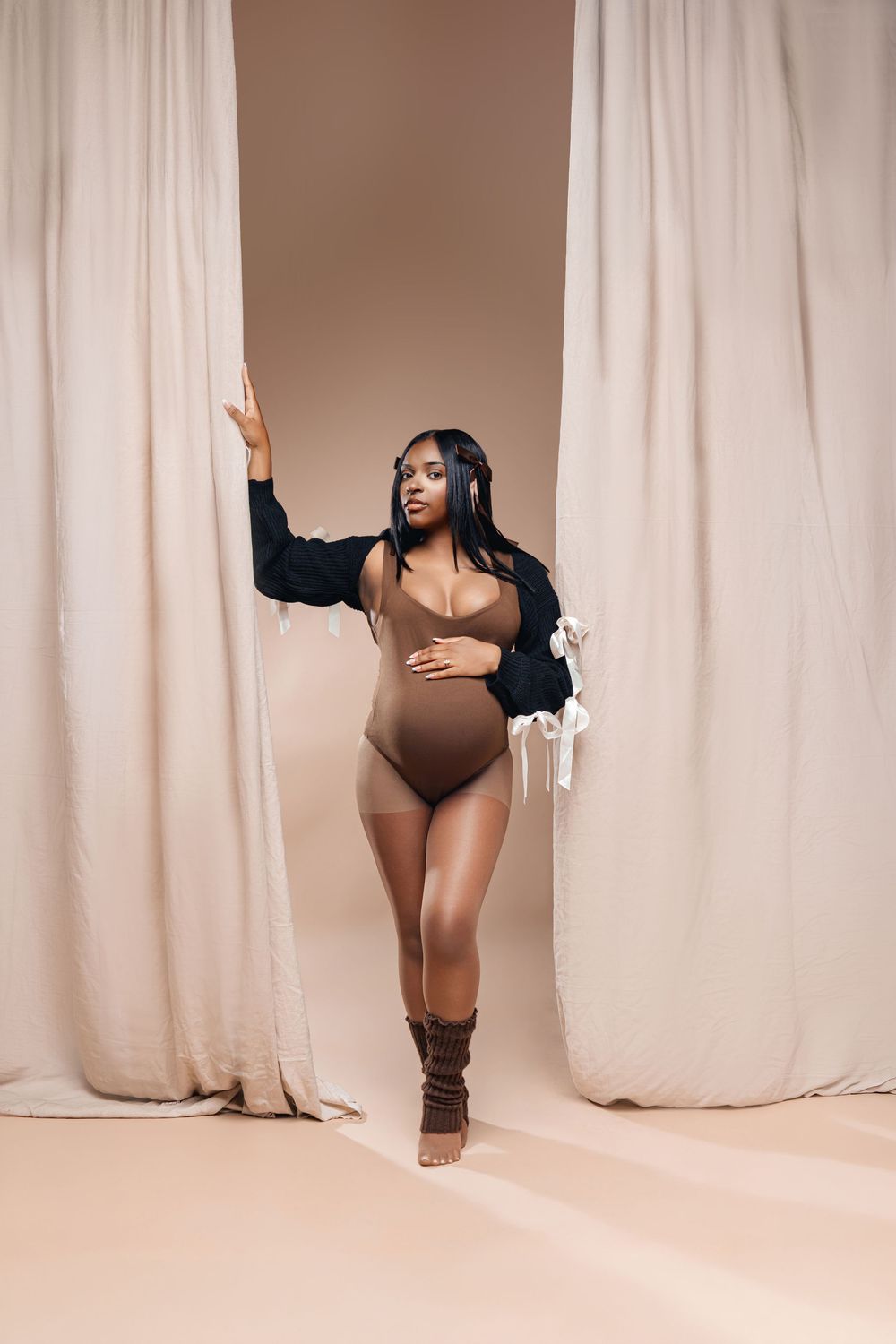 Pregnant Black woman in brown bodysuit and boots poses between beige curtains in a studio setting.