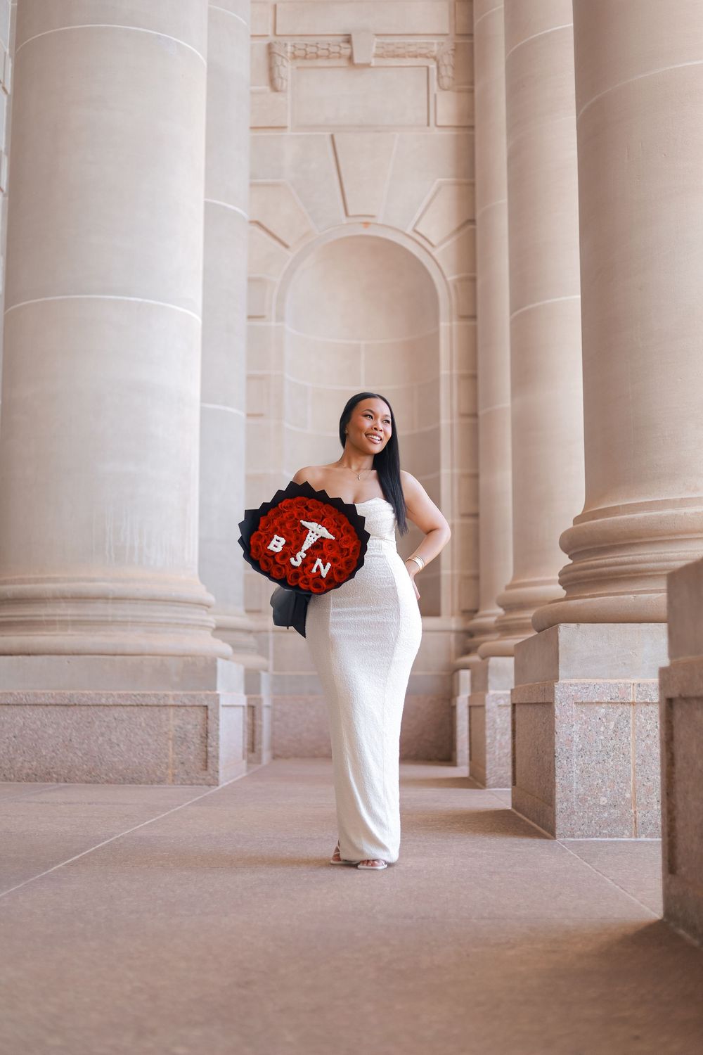 Woman in white gown holding red rose bouquet with BSN letters between elegant stone columns.
