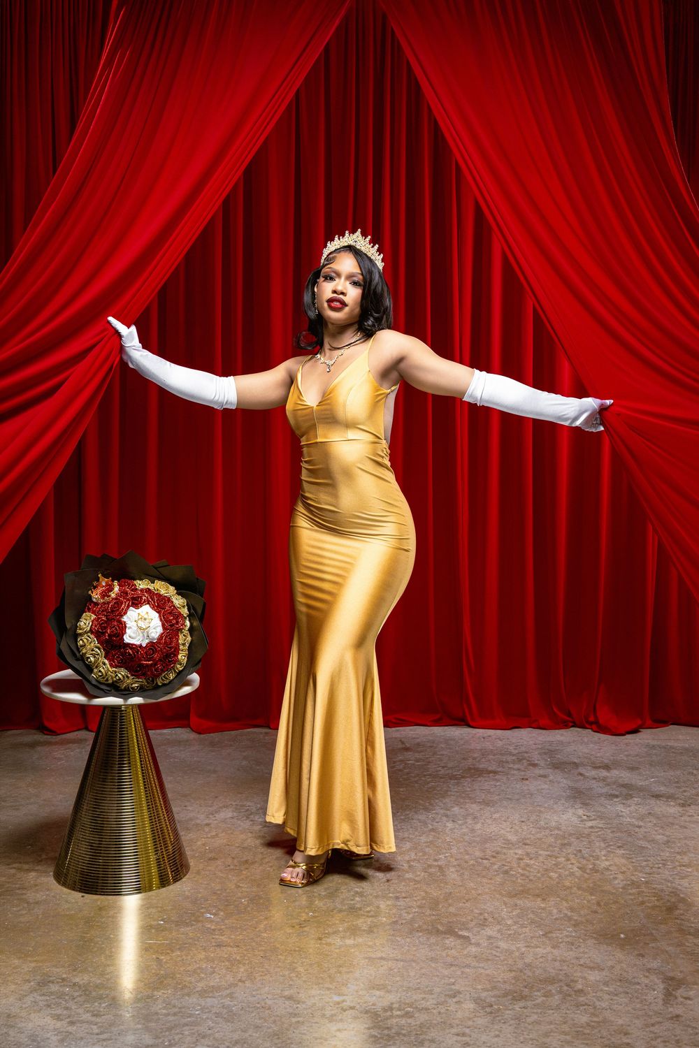 Woman in gold gown and crown poses gracefully before red curtains on stage.