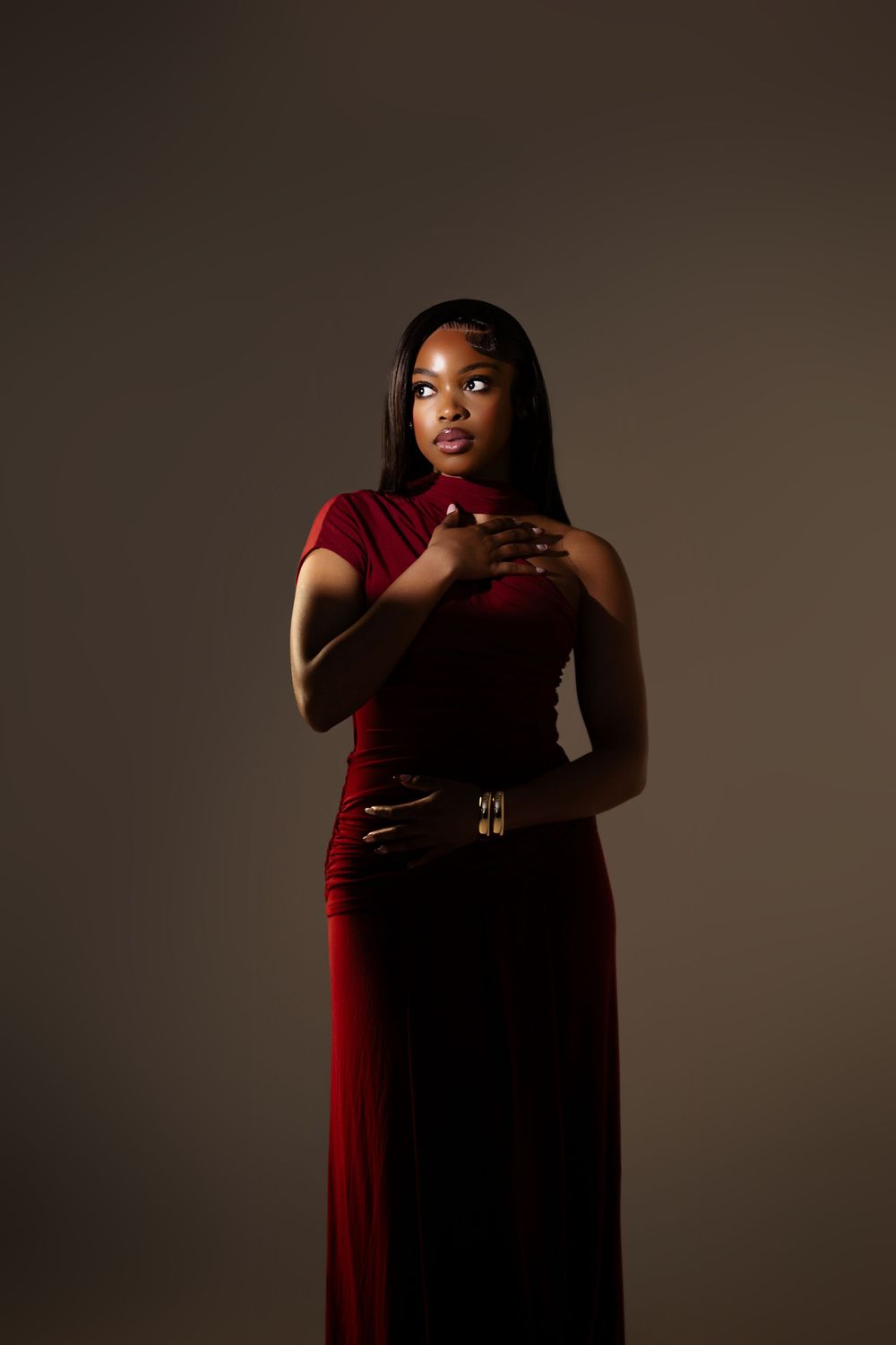A woman in a red velvet gown with gold belt poses elegantly against a dark studio background.