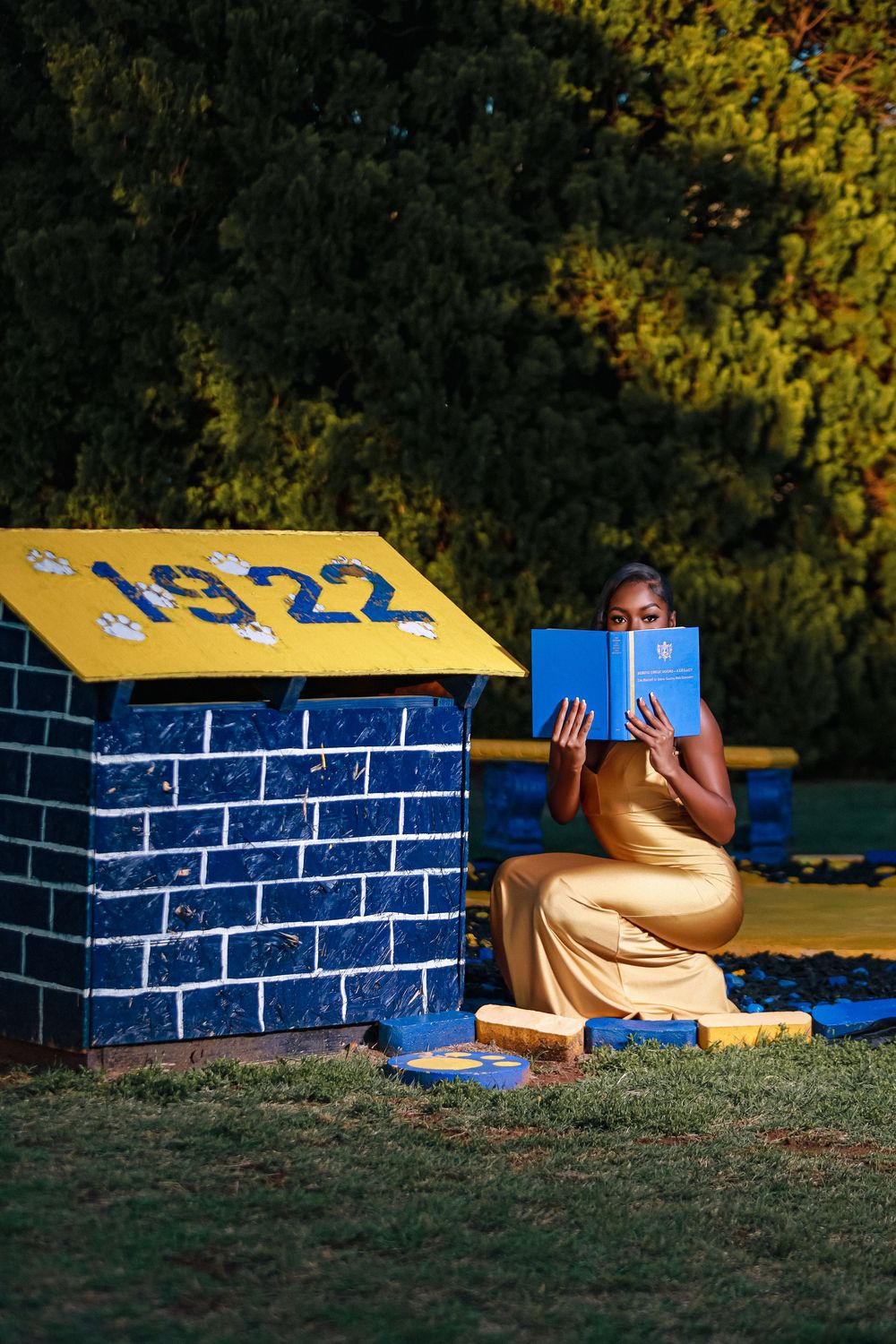 Woman in yellow dress reading a blue book beside a decorative blue and yellow brick structure with 1922 sign.