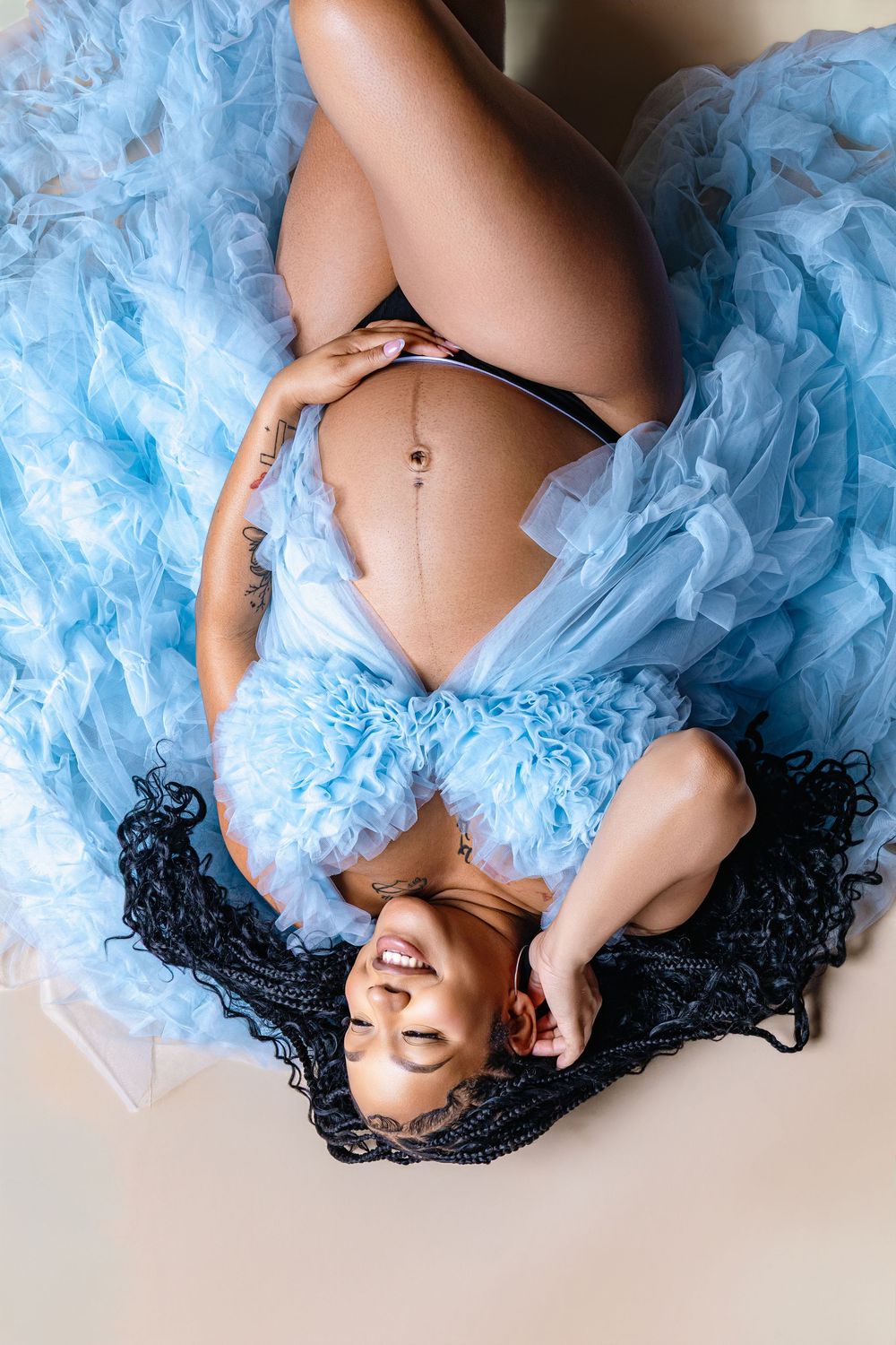 Pregnant woman in blue tulle robe lying down, smiling, with braided hair and visible baby bump.