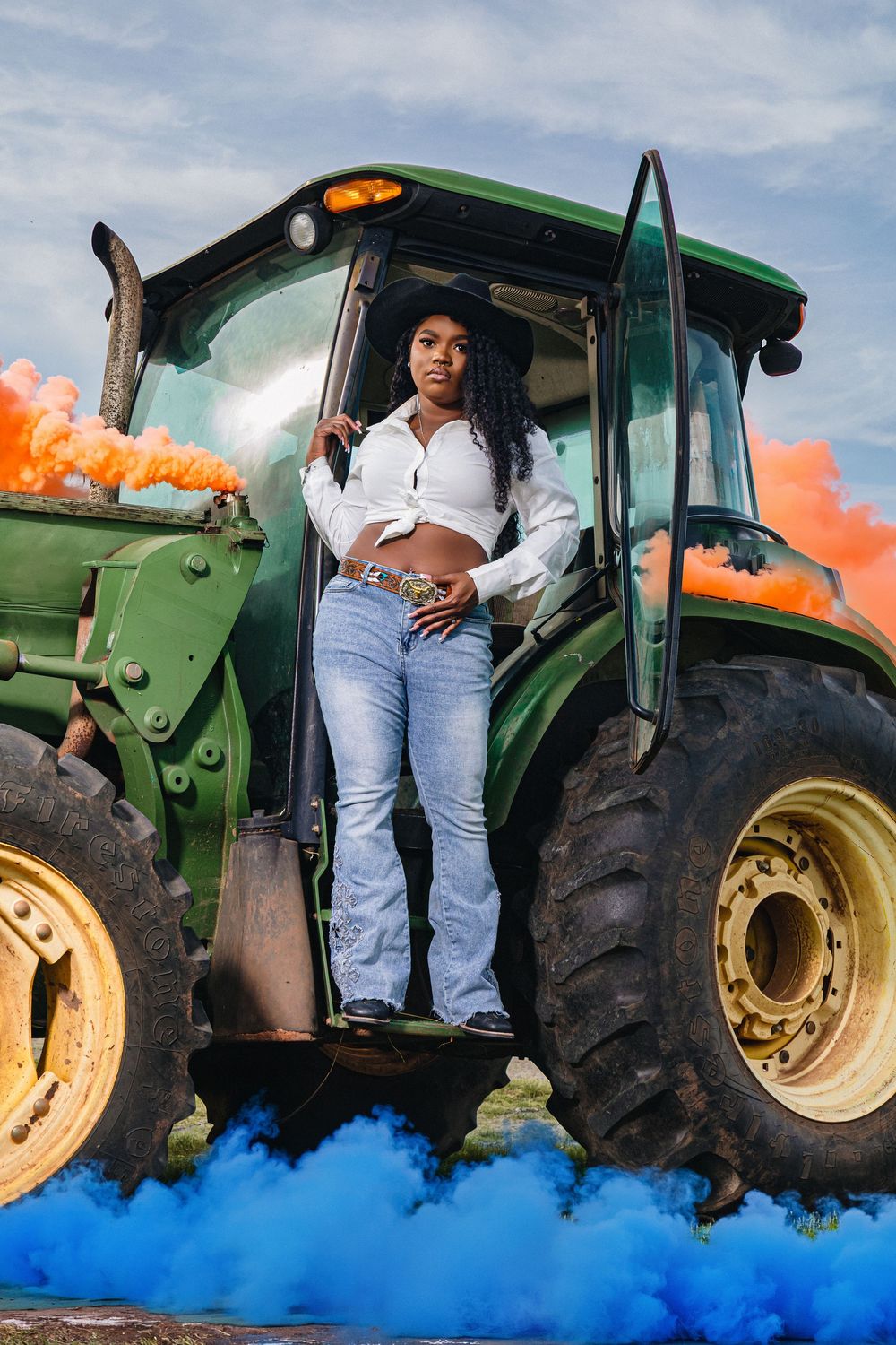 Young woman in white crop top and jeans poses on green tractor surrounded by vibrant orange and blue smoke.
