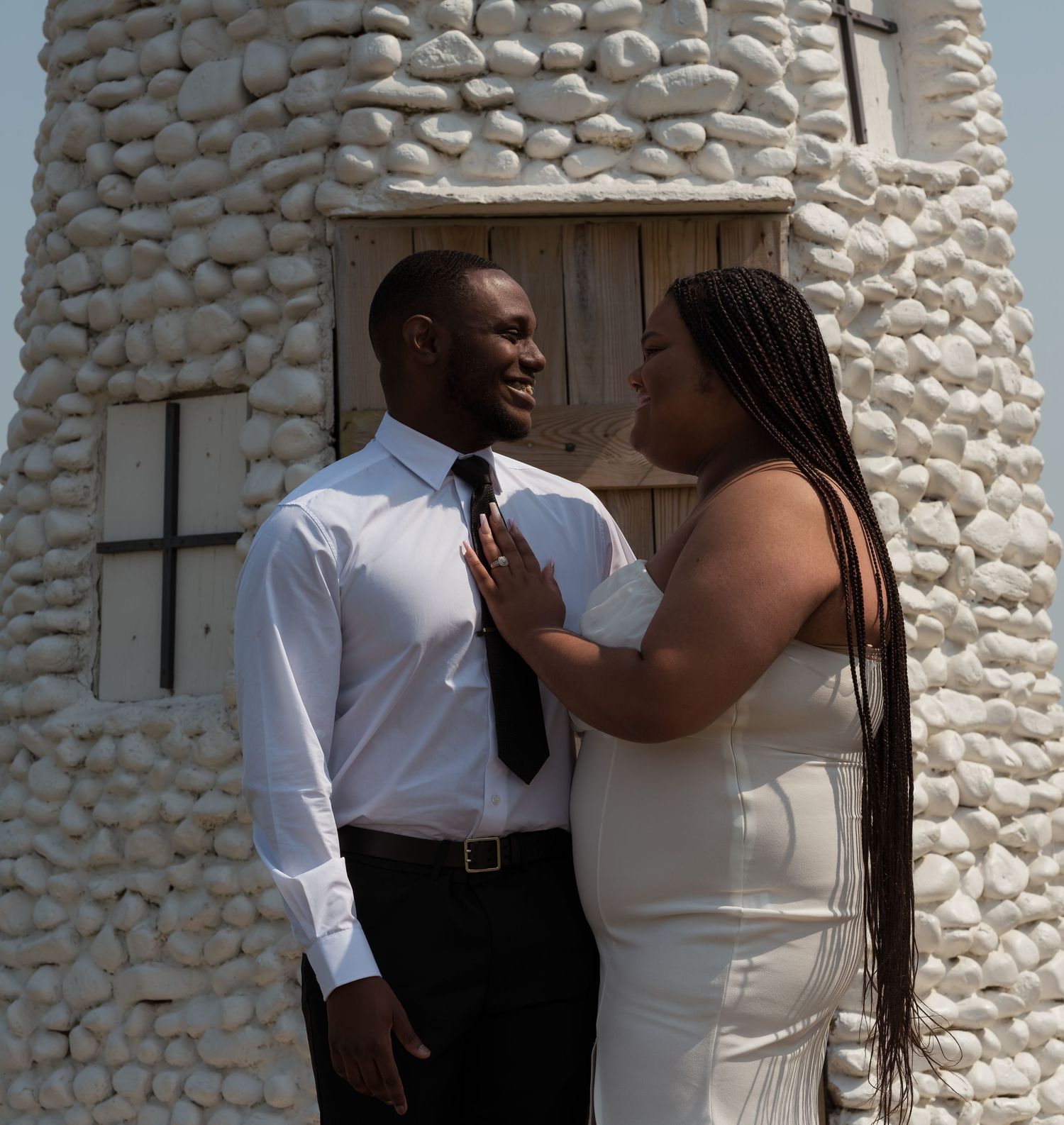 A romantic couple embraces tenderly against a white stone wall wearing formal wedding attire.