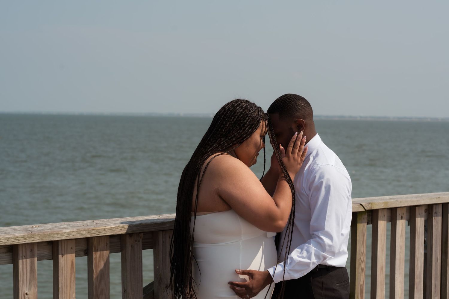 Couple shares an intimate kiss while standing on a wooden deck overlooking the ocean on a cloudy day.