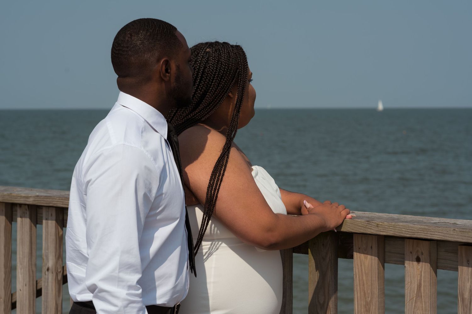 A couple on a wooden pier overlooks the ocean, with a sailboat visible on the horizon beneath a hazy sky.