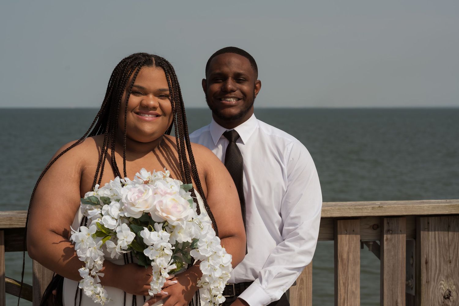 A joyful couple stands together on an oceanfront deck during their wedding celebration with white floral bouquet.