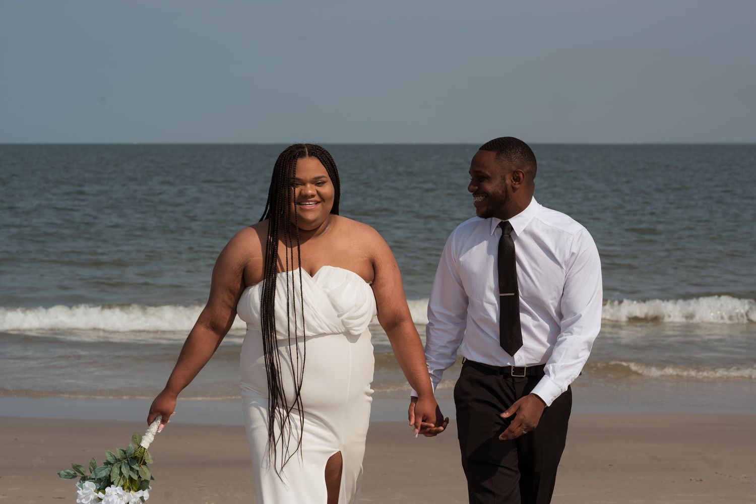 A couple walks hand in hand along the beach in formal attire.
