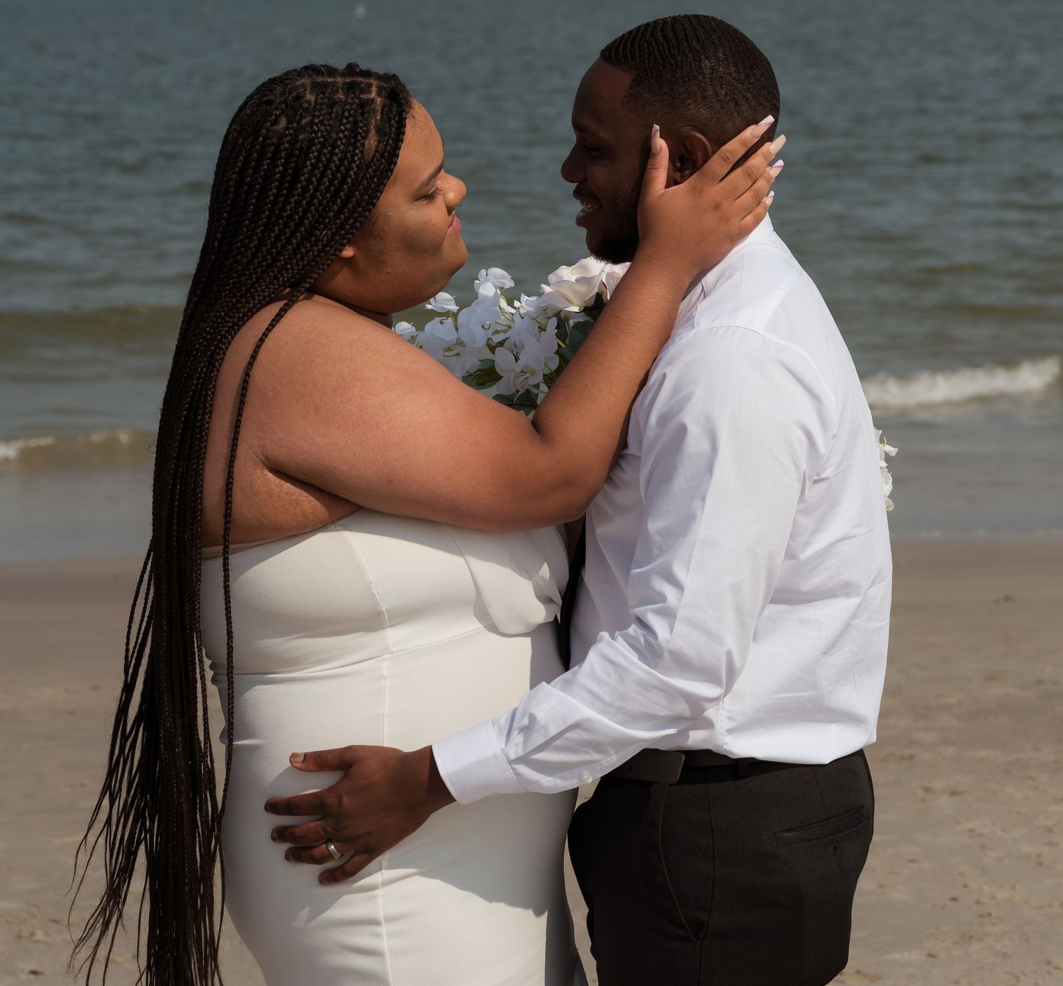 A loving couple embraces on a sandy beach during what appears to be a wedding photoshoot.