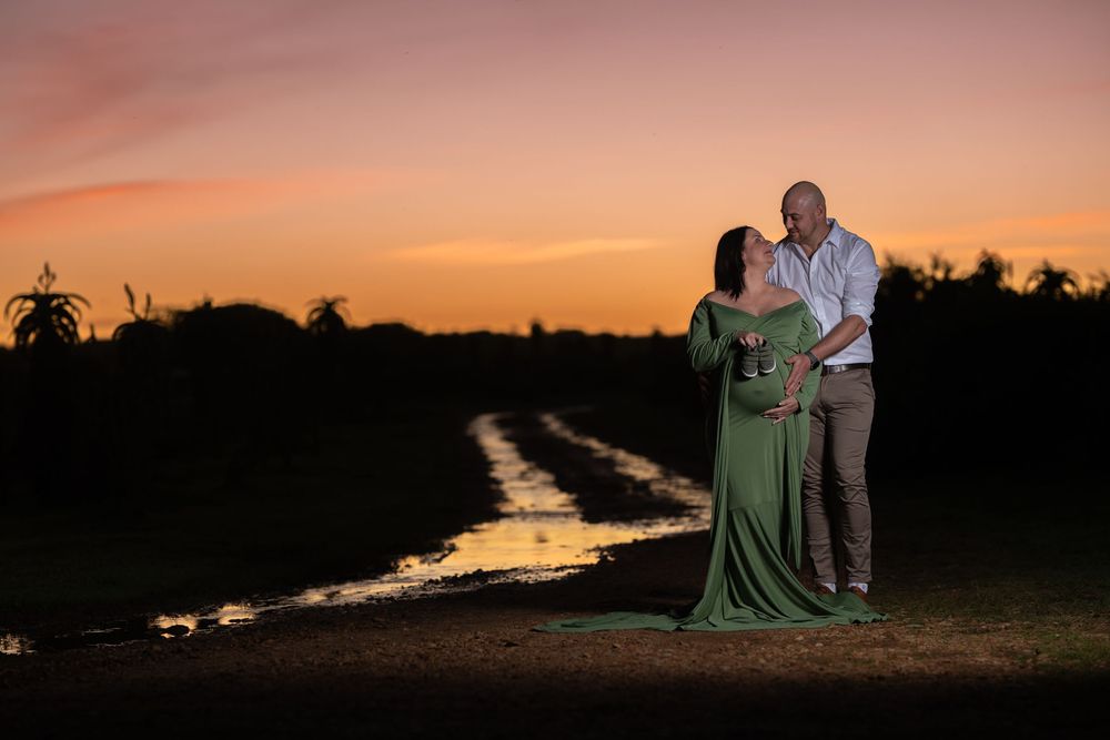 Maternity couple embracing at vibrant orange sunset beside reflective water path in open field.