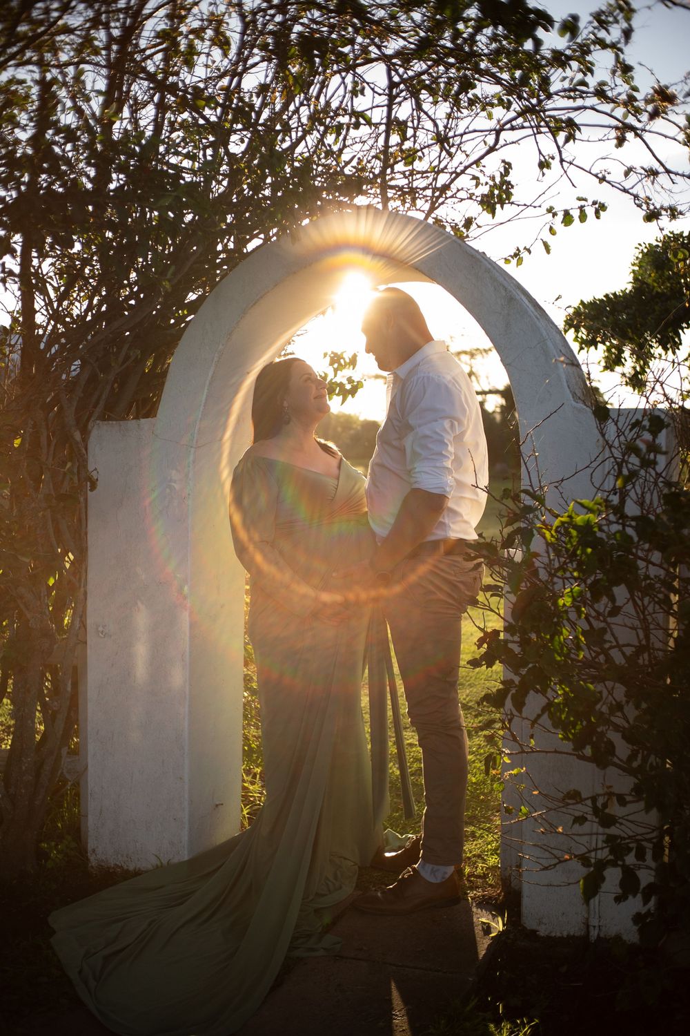 Couple standing under white arch with golden sun flare, woman in flowing green maternity gown.