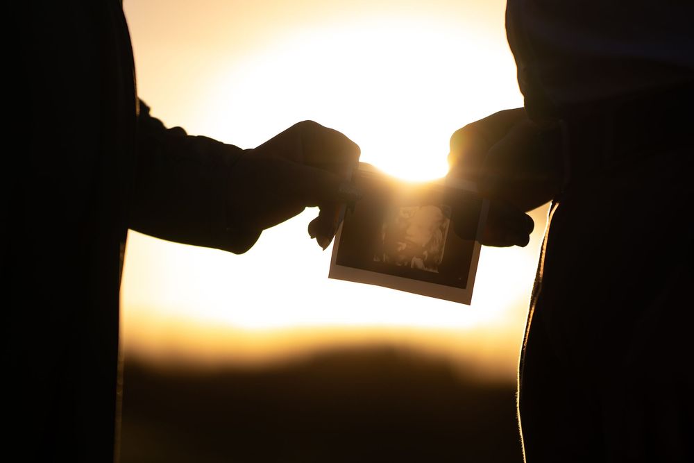 Silhouette of two people exchanging a photograph against a warm golden sunset background.