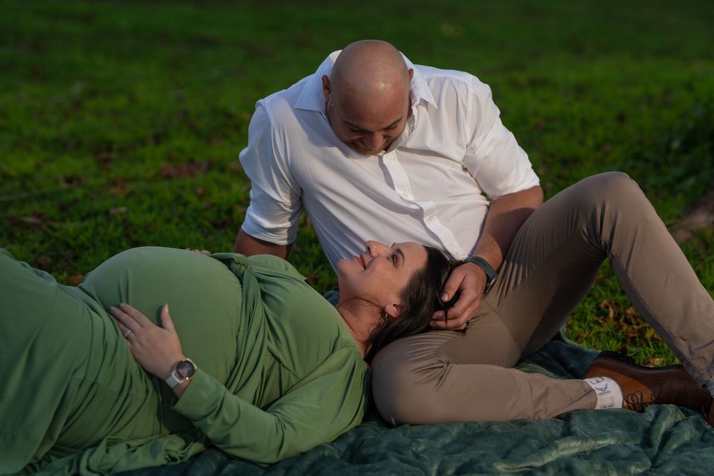 Expectant couple relaxing on grass, woman in green dress lying down as partner gazes lovingly.