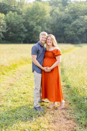 Expecting couple poses together in field wearing blue shirt and orange dress in Springfield, Missouri.