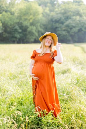 Pregnant woman in orange maxi dress and sun hat poses joyfully in summer field in Springfield, Missouri.