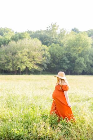Pregnant woman in orange dress and hat walks through golden field at sunset in Springfield, Missouri