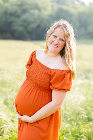 Pregnant woman in orange off-shoulder dress smiles warmly in sunlit field in Springfield, Missouri.