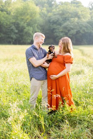 Couple holding small dog poses together in sunny field during maternity photoshoot in Springfield, Missouri