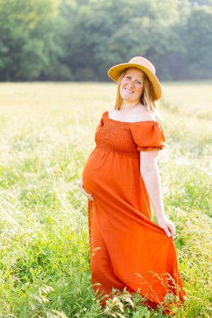 Expectant mother in flowing orange dress and straw hat stands in sunny meadow in Springfield, Missouri.