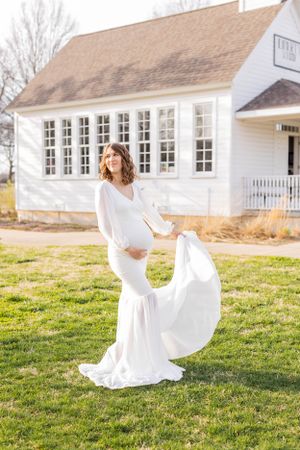 Maternity photo of woman in flowing white dress standing on grass in front of white building in Springfield, Missouri.