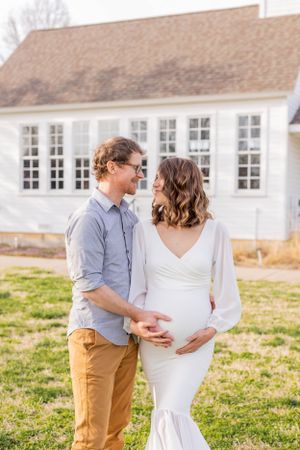 Expecting couple stands together in front of white farmhouse during maternity photoshoot in Springfield, Missouri.