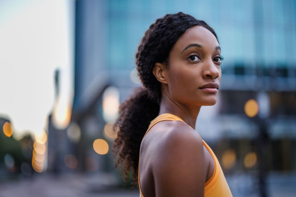 A young Black woman in a yellow tank top with braided ponytail poses outdoors against a blurred urban cityscape backdrop.