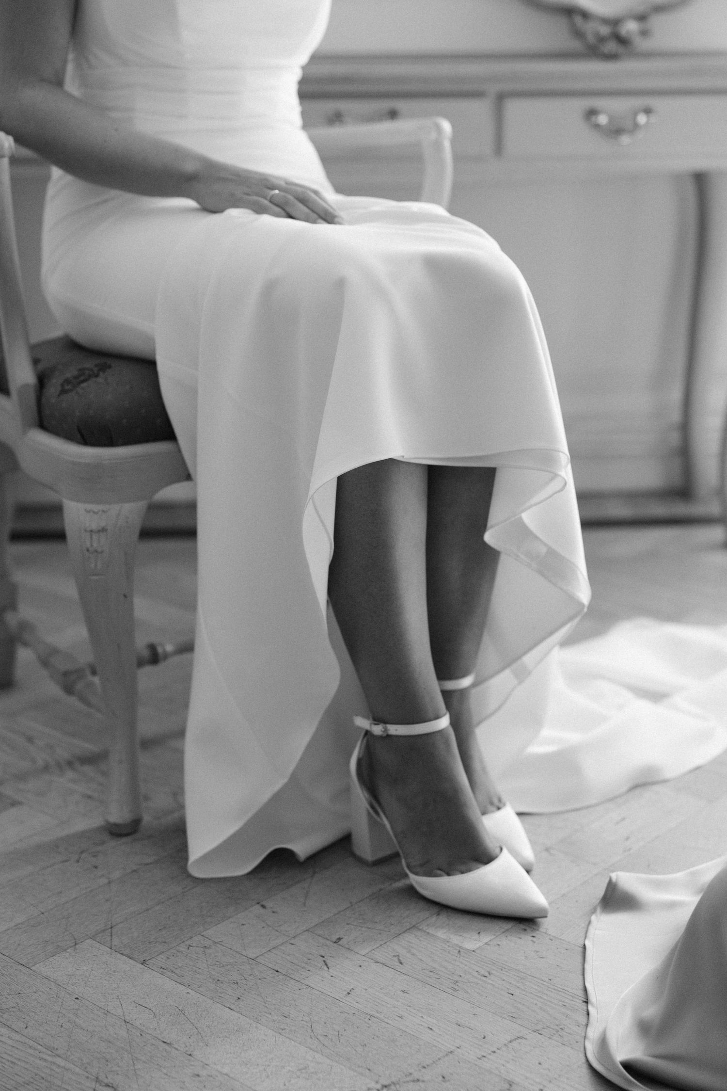 Close-up black and white photo of white bridal shoes and flowing dress hem on wooden floor.