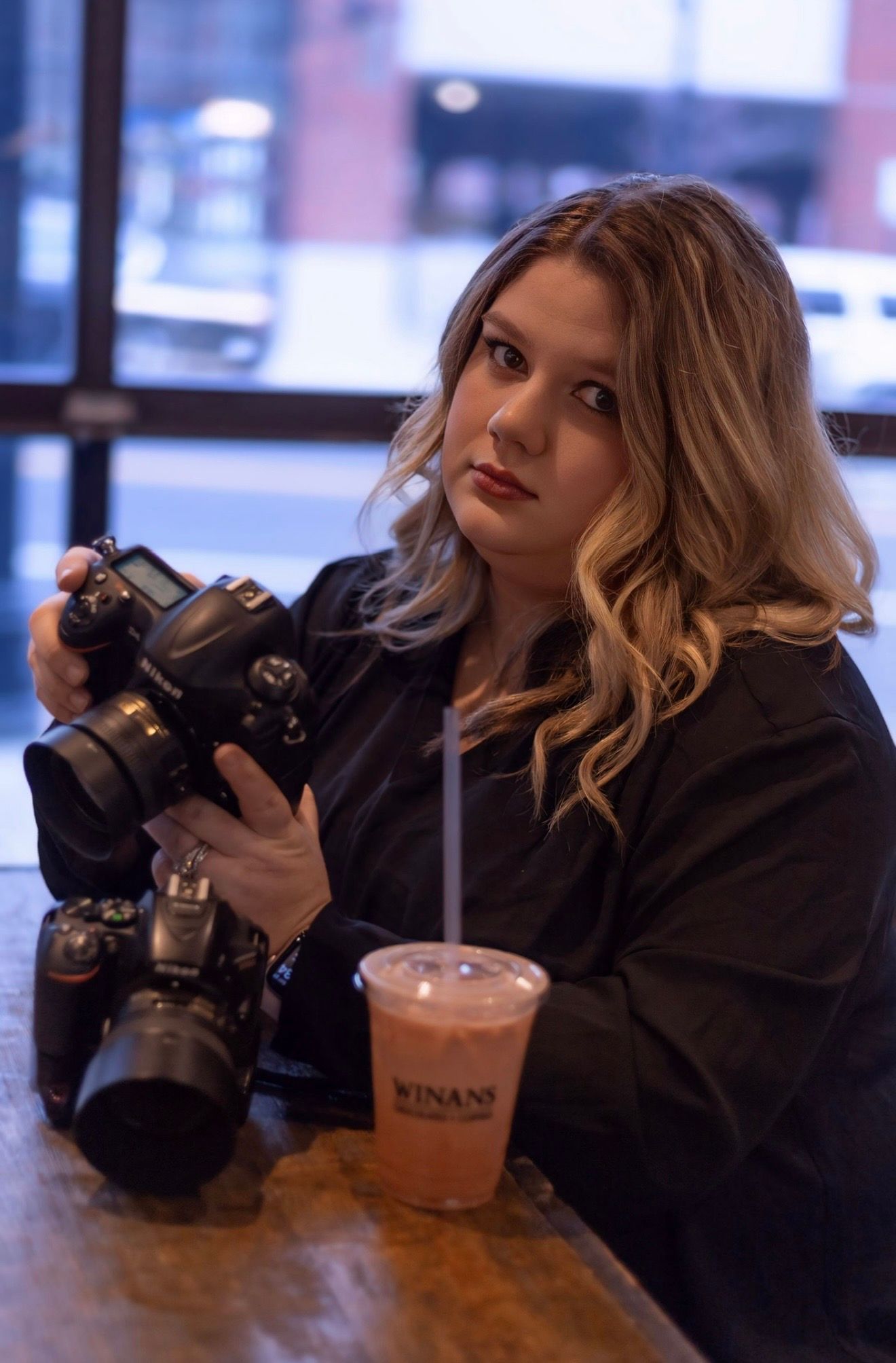 Blonde female photographer holding a Nikon camera at a cafe table with a drink nearby.