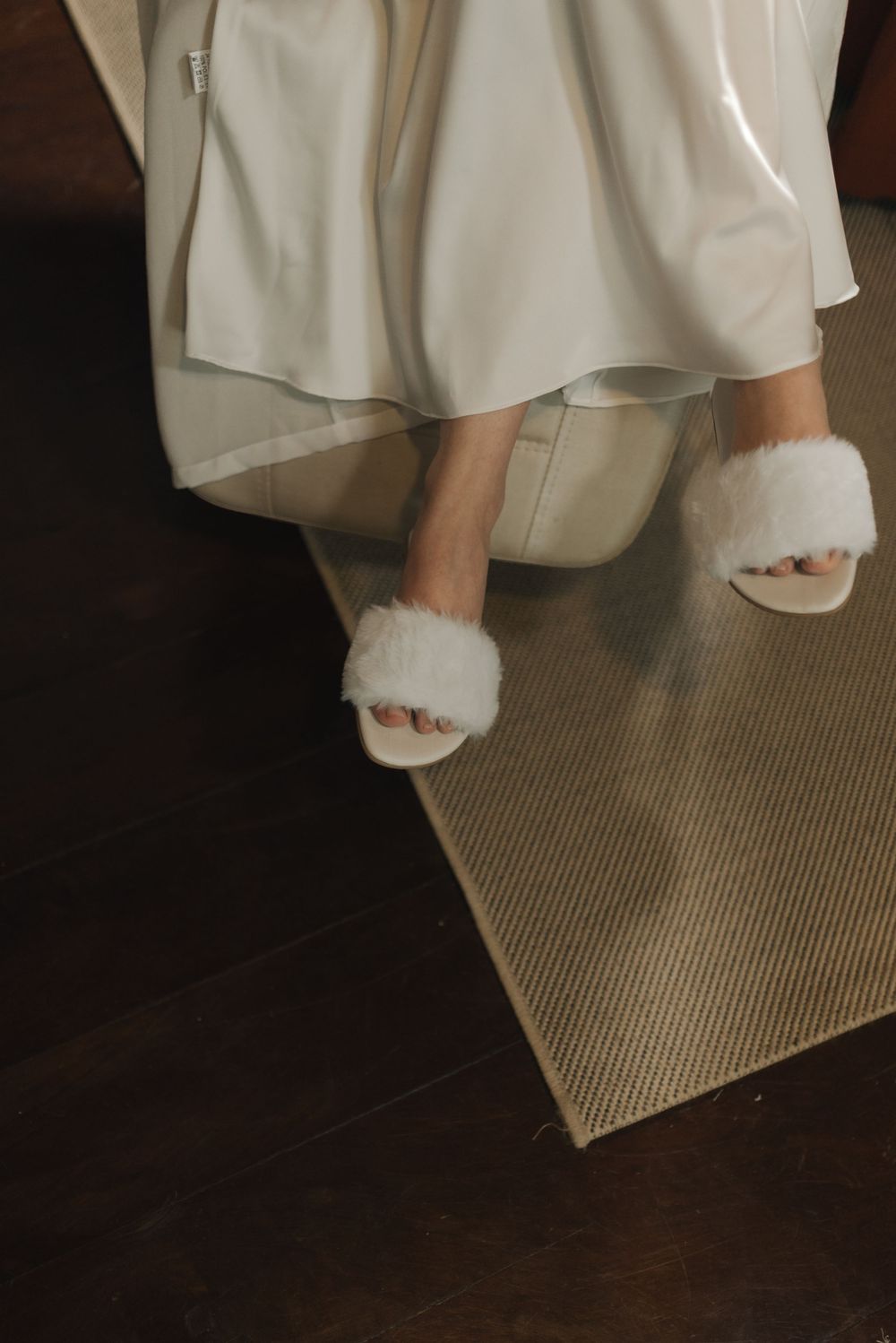 Cozy white fluffy slippers peek out beneath a flowing ivory dress hem on a textured carpet.