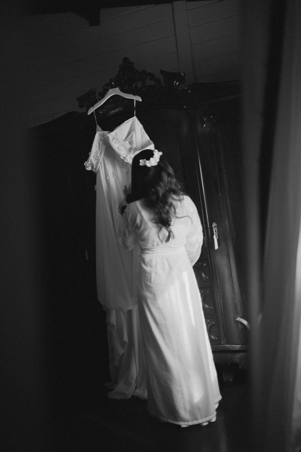 A black and white photograph shows a flowing white wedding dress hanging on display in a dark room.