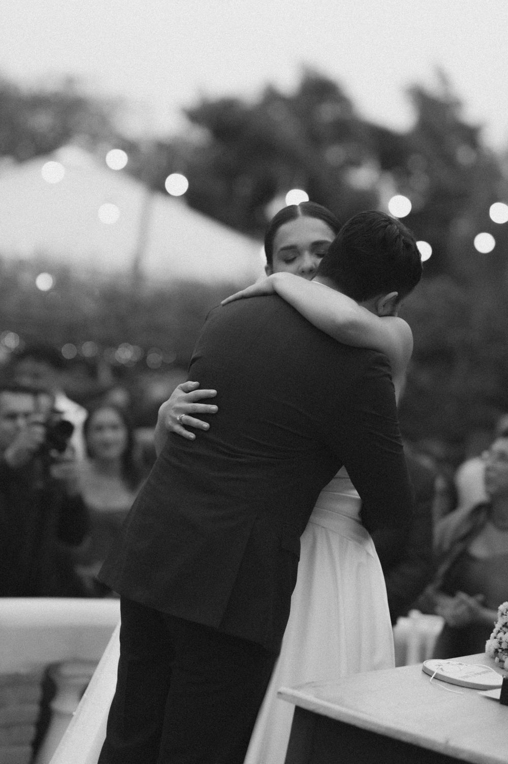 A tender embrace between a couple in formal attire dancing at an outdoor wedding reception with twinkling lights in background.