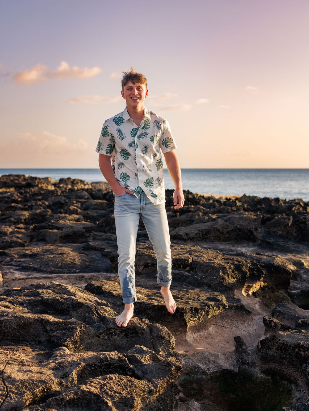 Young man in tropical shirt and jeans stands barefoot on rocky shore at sunset with ocean in background.