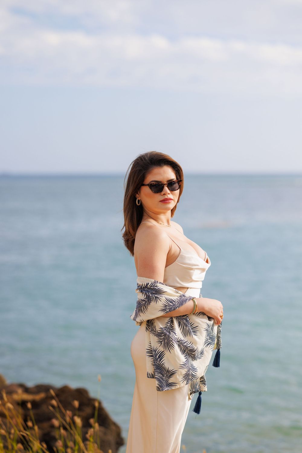 Woman in cream dress and sunglasses poses by ocean shoreline with floral shawl on sunny day.