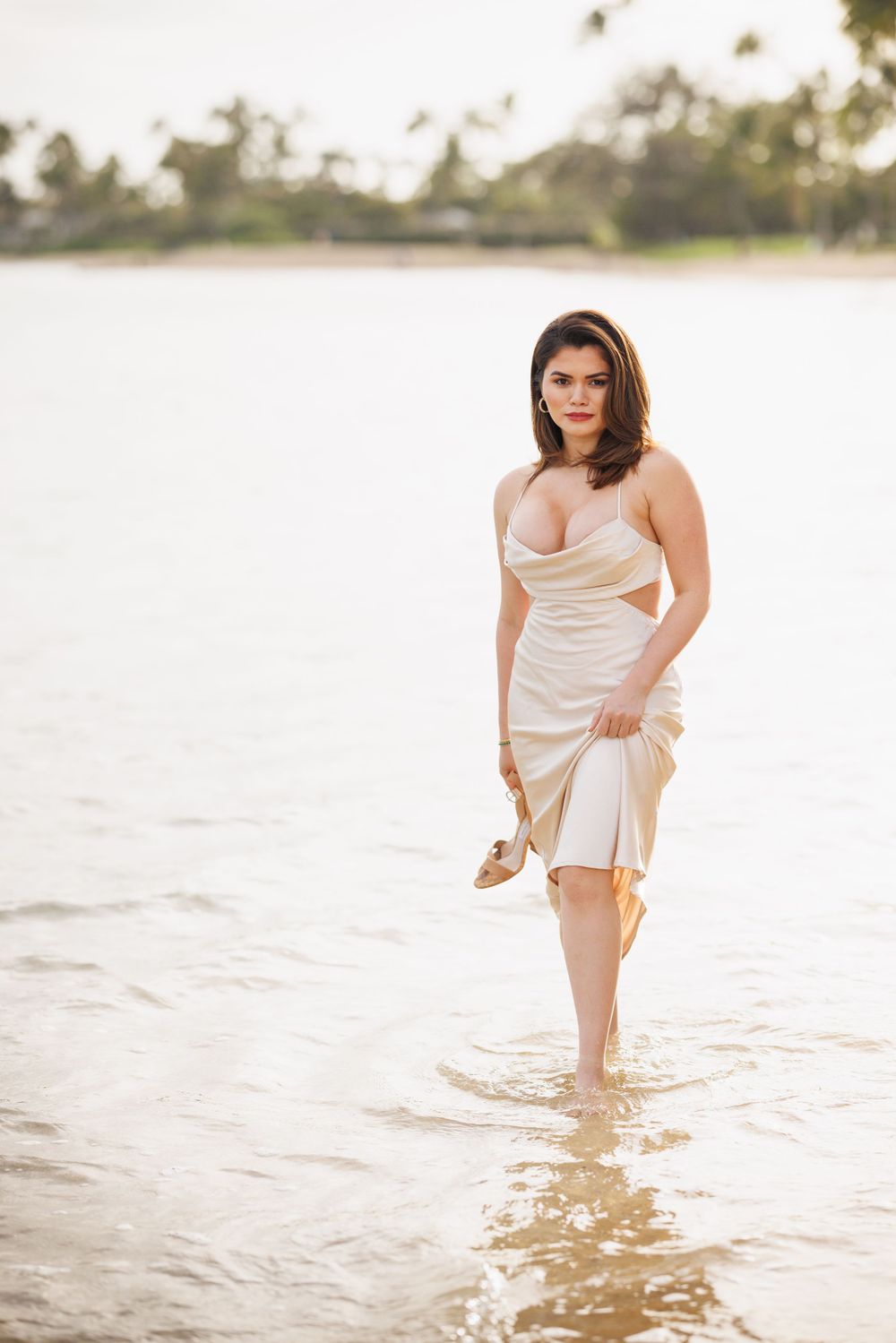 Woman in elegant cream dress wading in shallow water, holding heels, with tropical palm trees in background.