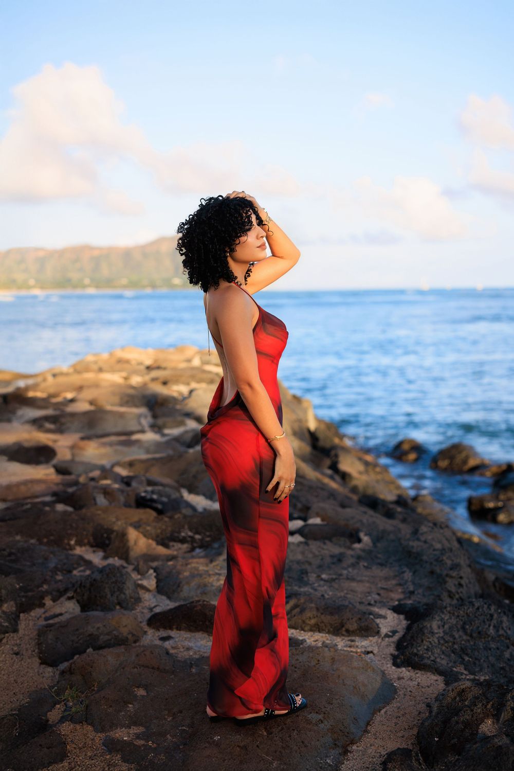 Woman in red dress standing on rocky shoreline with ocean and Diamond Head backdrop in golden hour light.