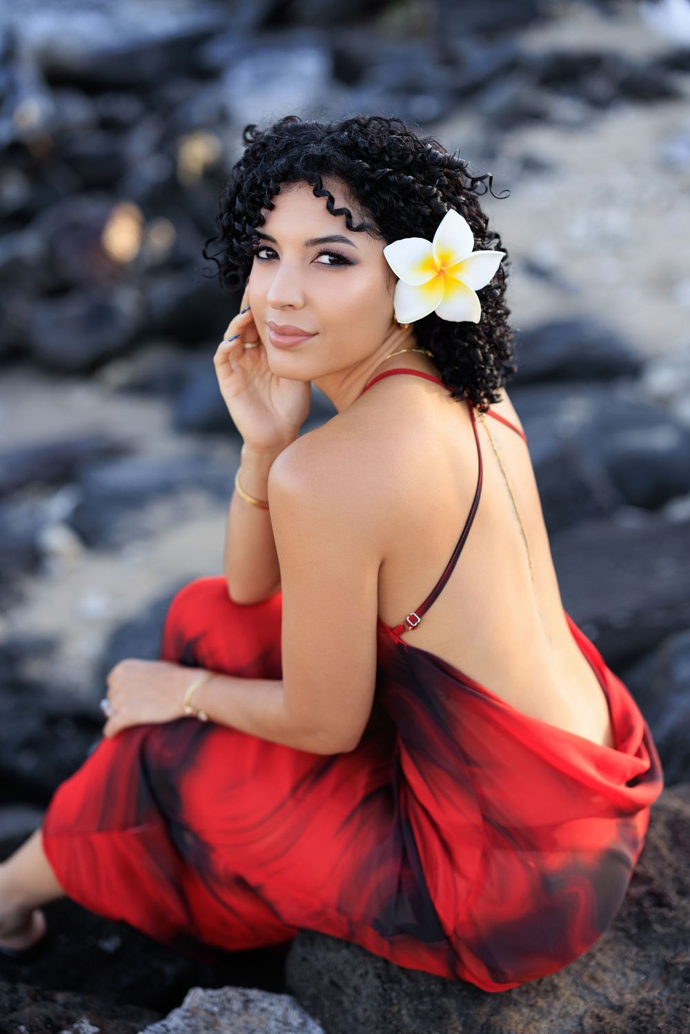Young woman in red backless dress with plumeria flower in curly hair poses on rocks in tropical setting.