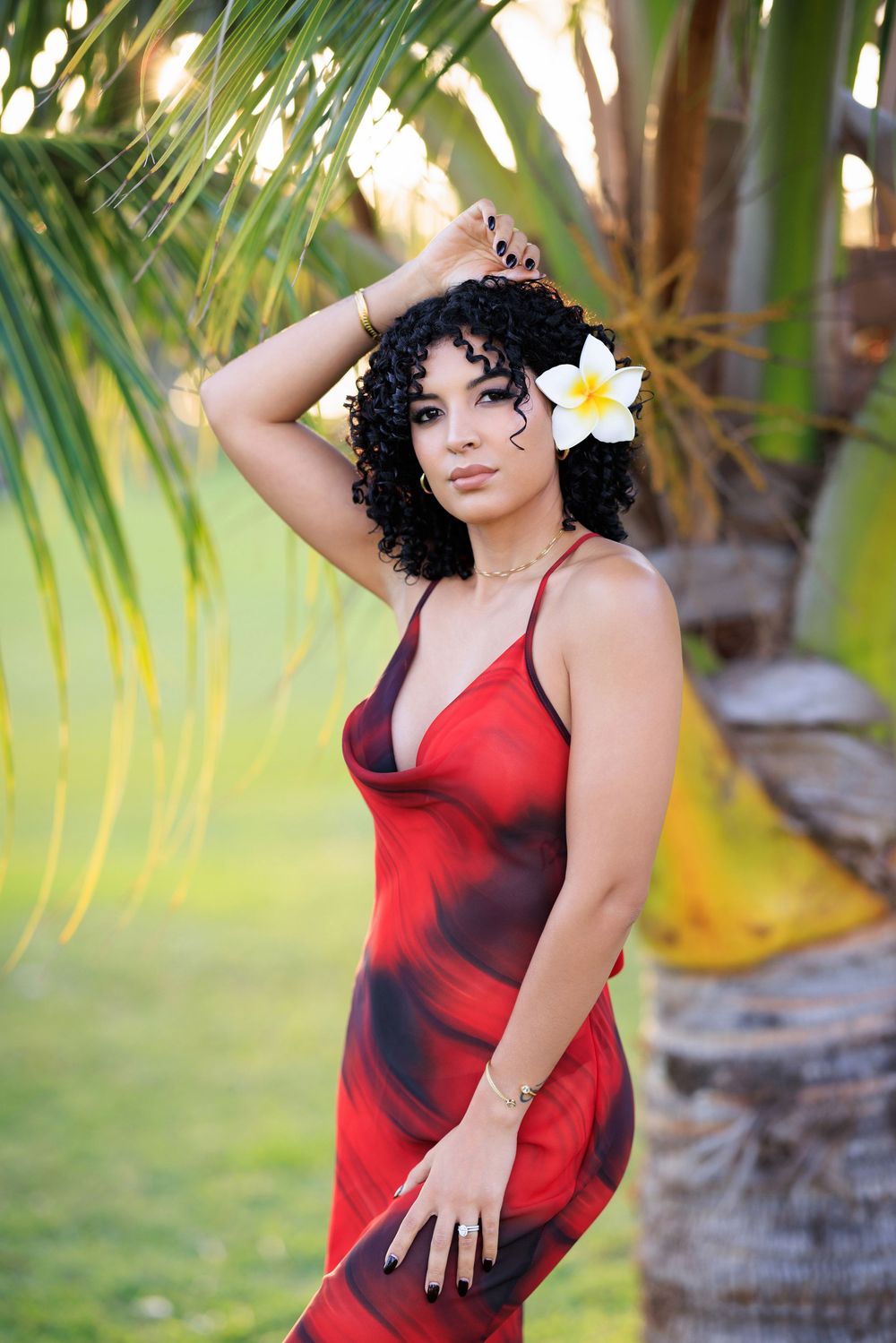 Woman in red dress with plumeria flower in curly hair posing near tropical palm tree outdoors.