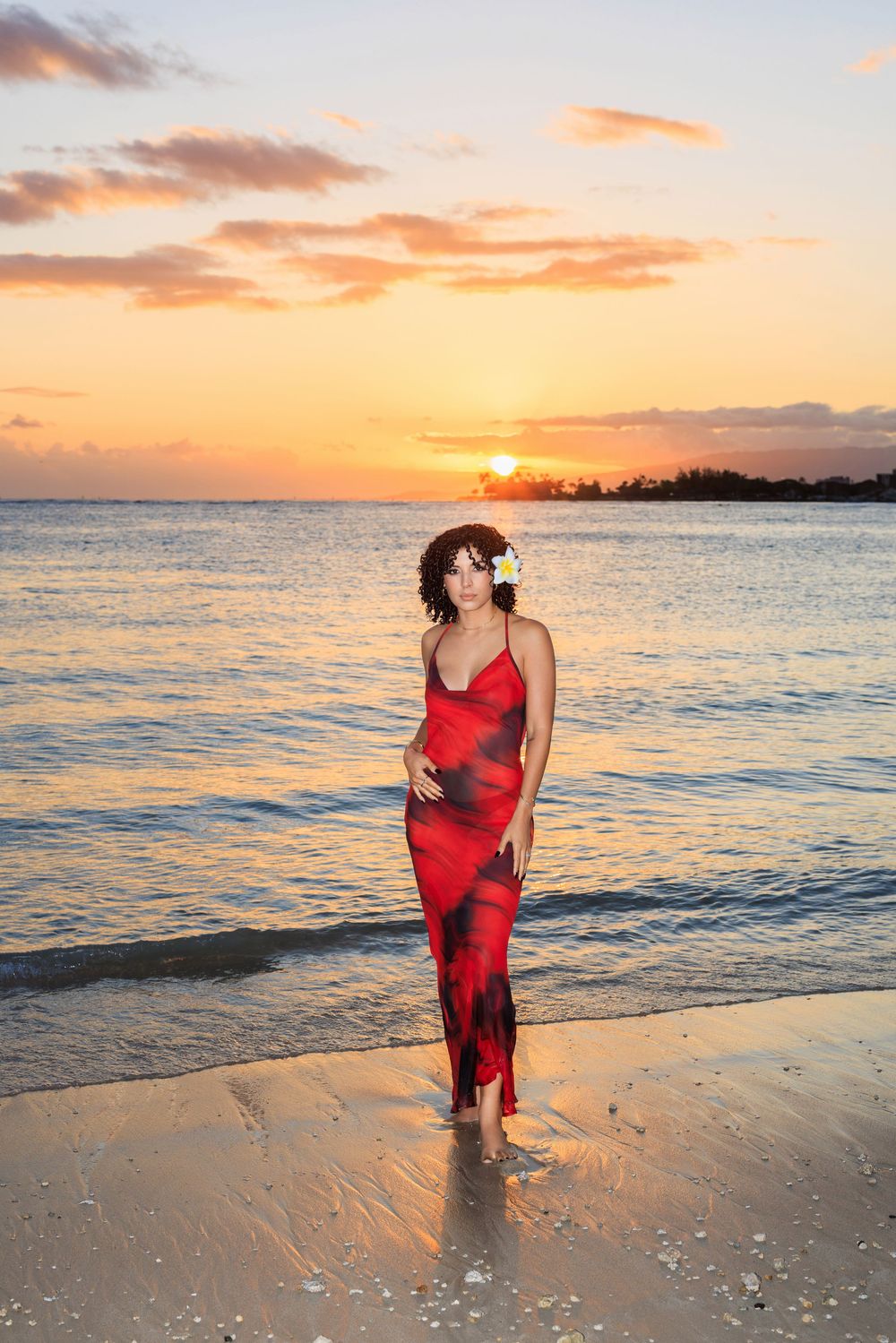 Woman in red dress with flower in hair stands on beach at golden sunset.