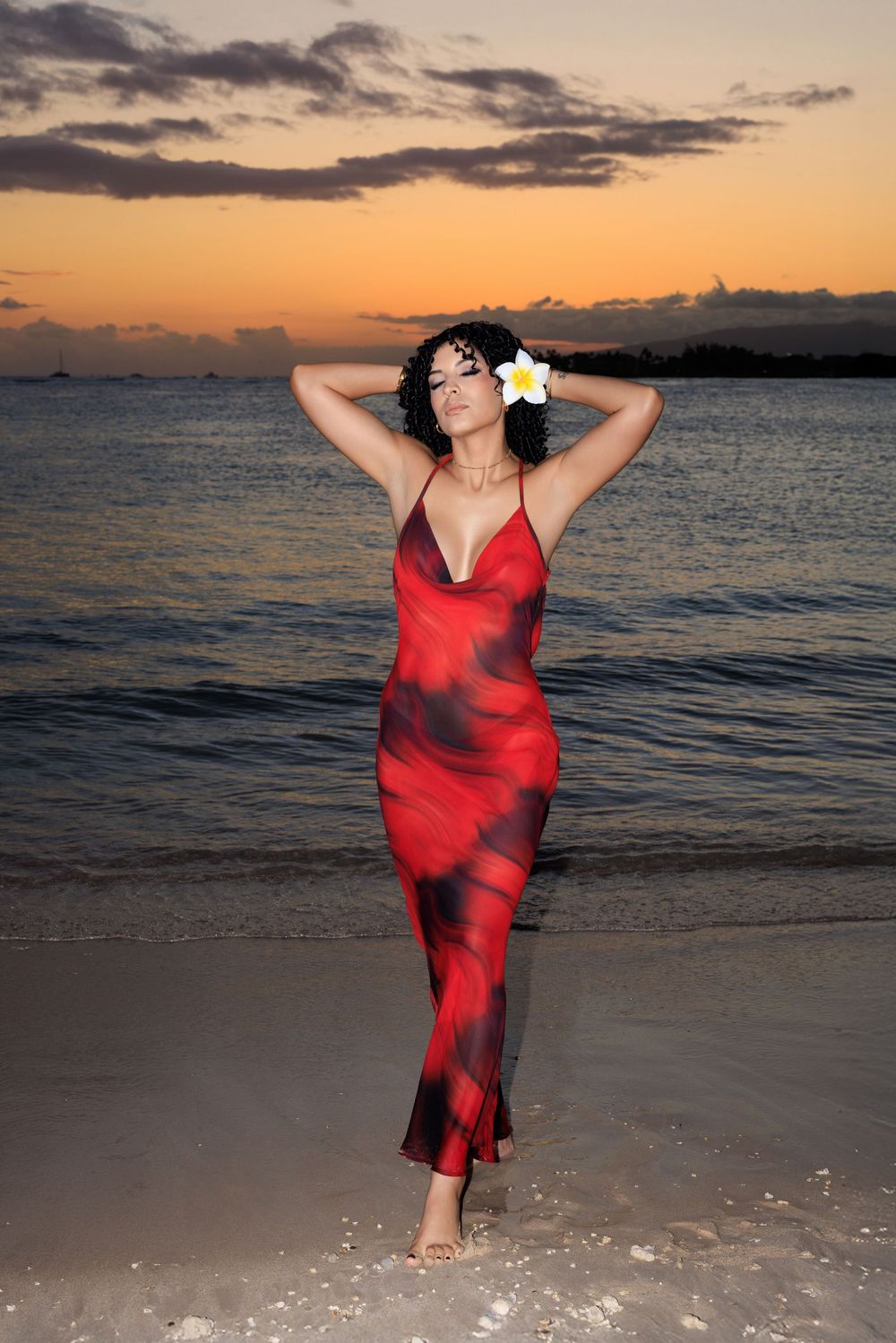 Woman in red floral maxi dress with plumeria flower in hair poses on tropical beach at sunset.
