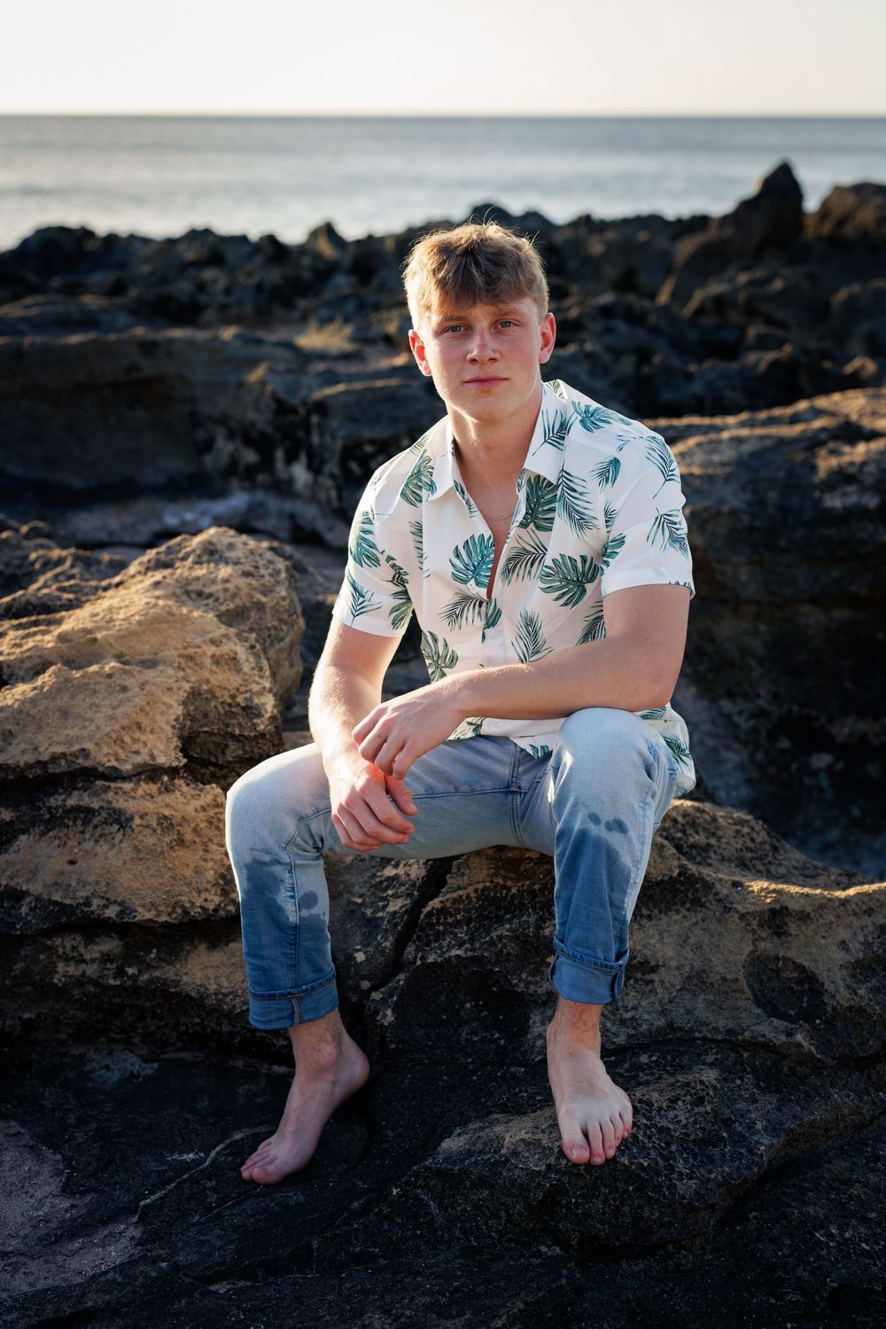 Young man in tropical shirt and jeans sitting barefoot on rocky shore with ocean horizon in background.