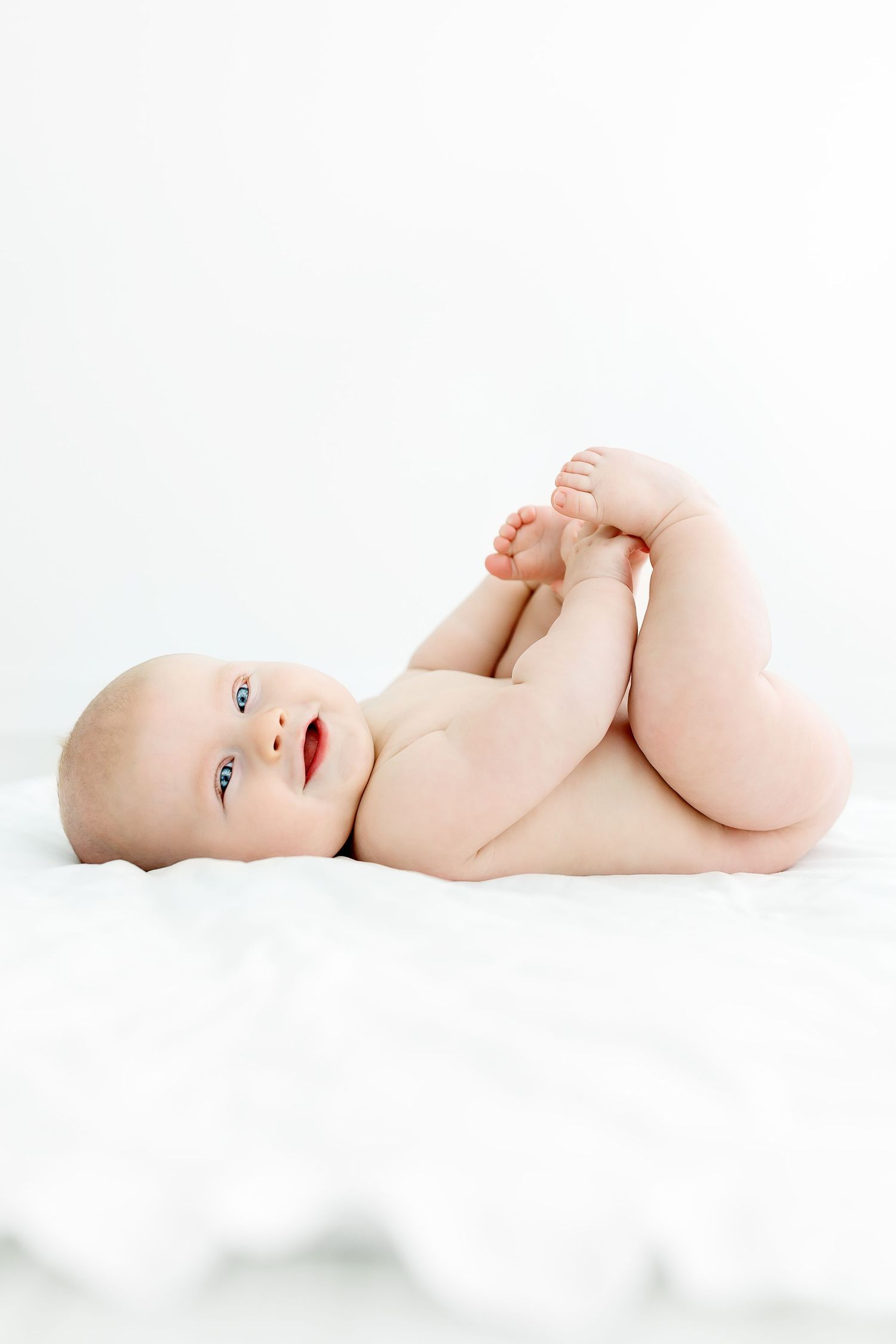 Sweet baby lying on white background holding their feet while looking up with bright eyes and soft skin.