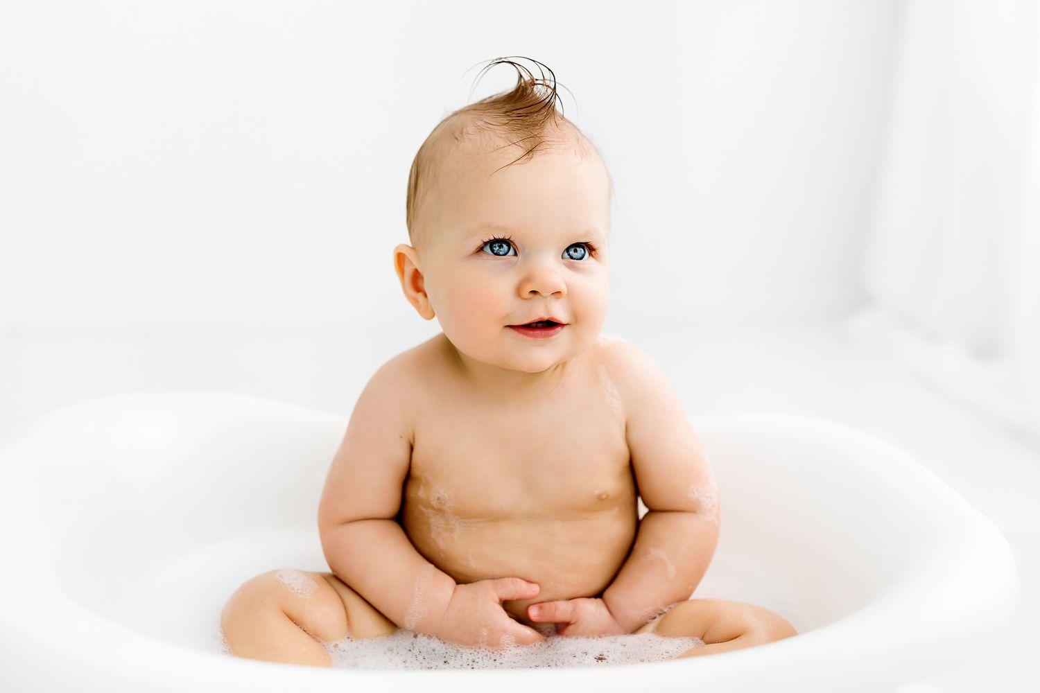Happy smiling baby with wispy hair sitting on white blanket against bright white background.