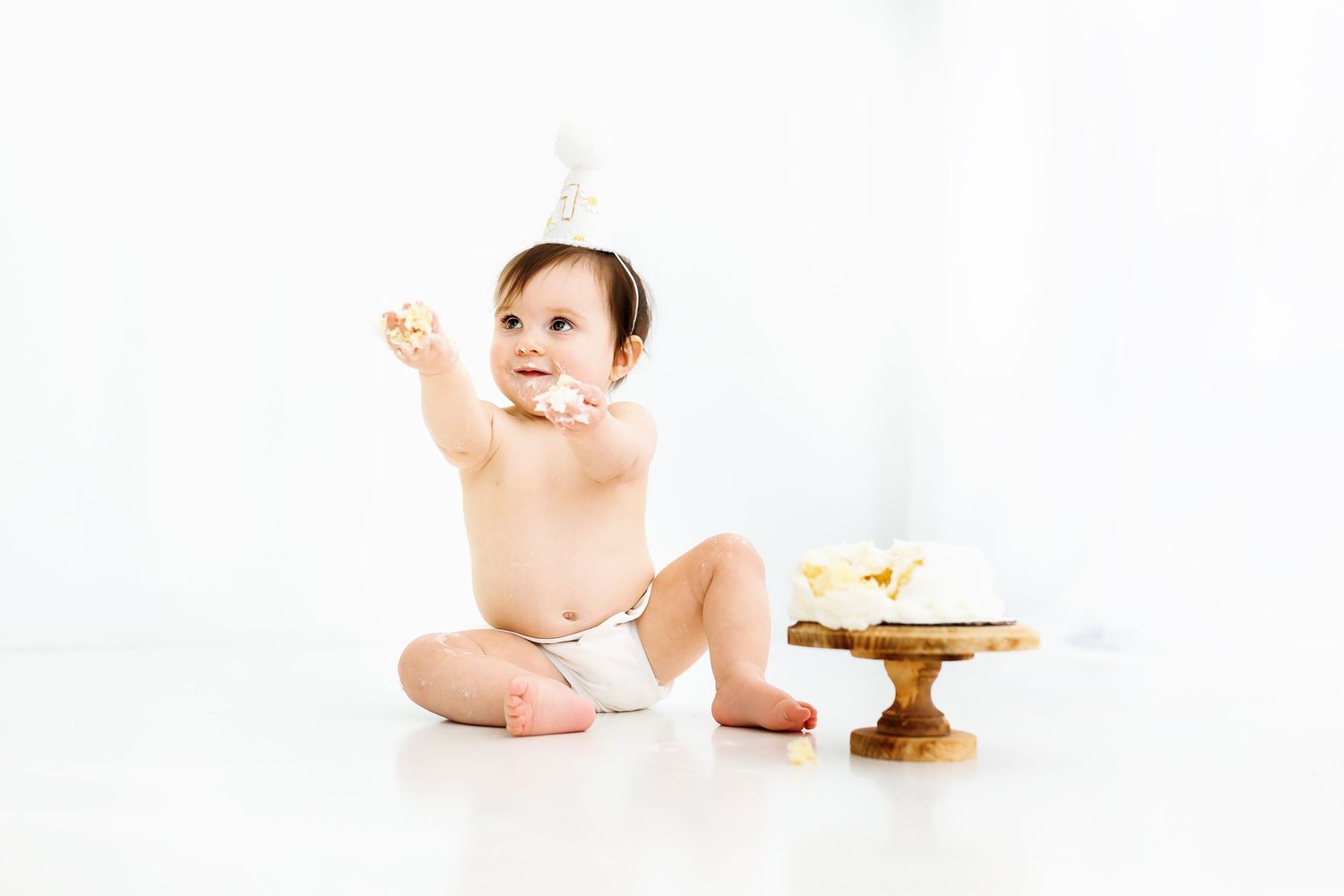 Small baby in white diaper sitting next to gold cake stand with white frosted cake in cake smash photo series.