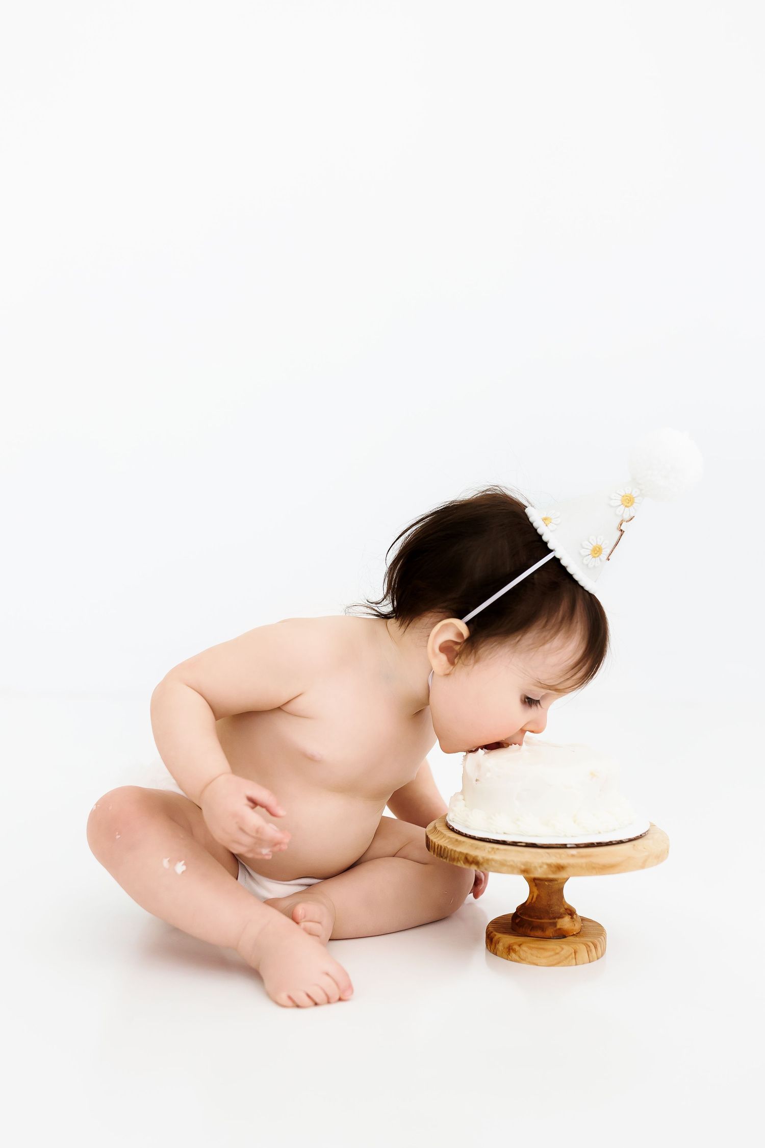 A baby in white diaper crawls and leans towards a small round cake on a wooden stand against white background.