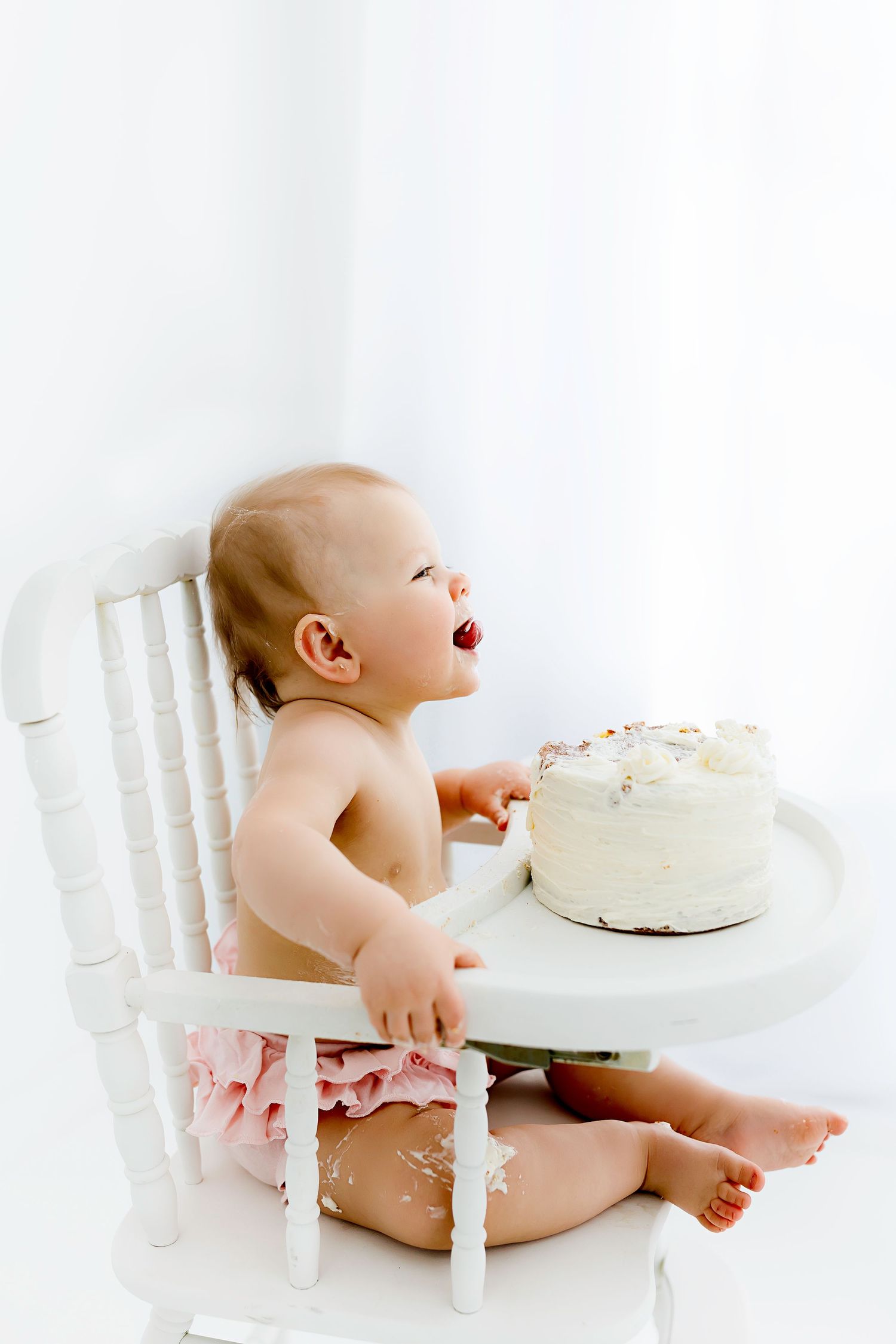 A baby sitting in a white chair reaches for a simple white frosted cake during a cake smash photo session.