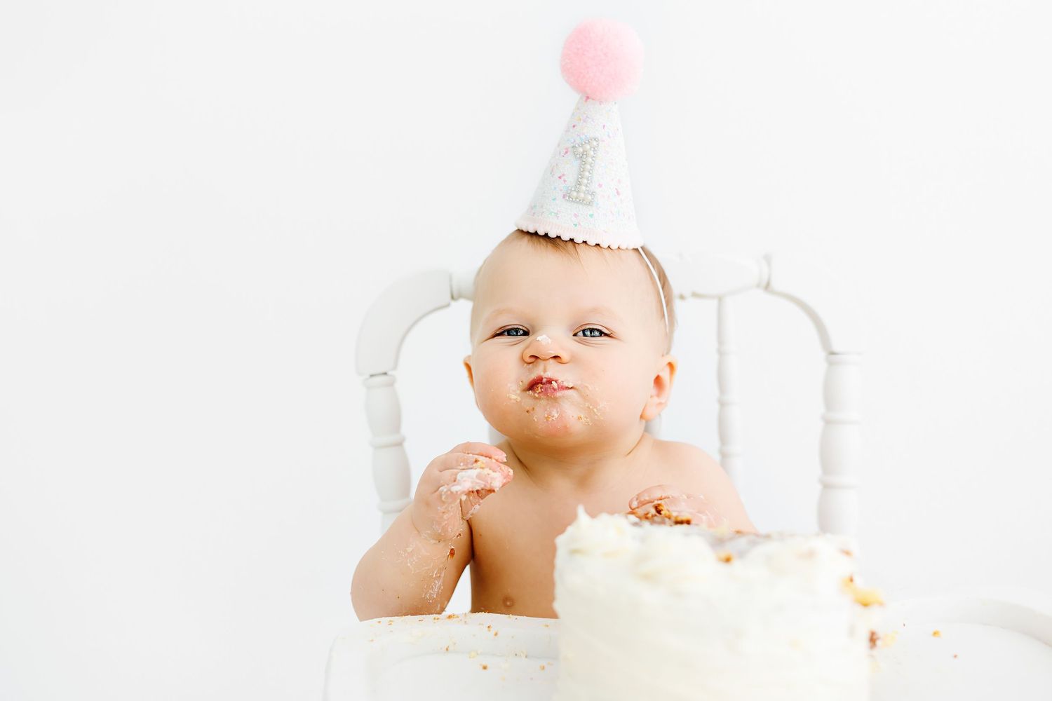 A baby drinks milk from a glass bottle while wearing a white outfit in a bright nursery setting.
