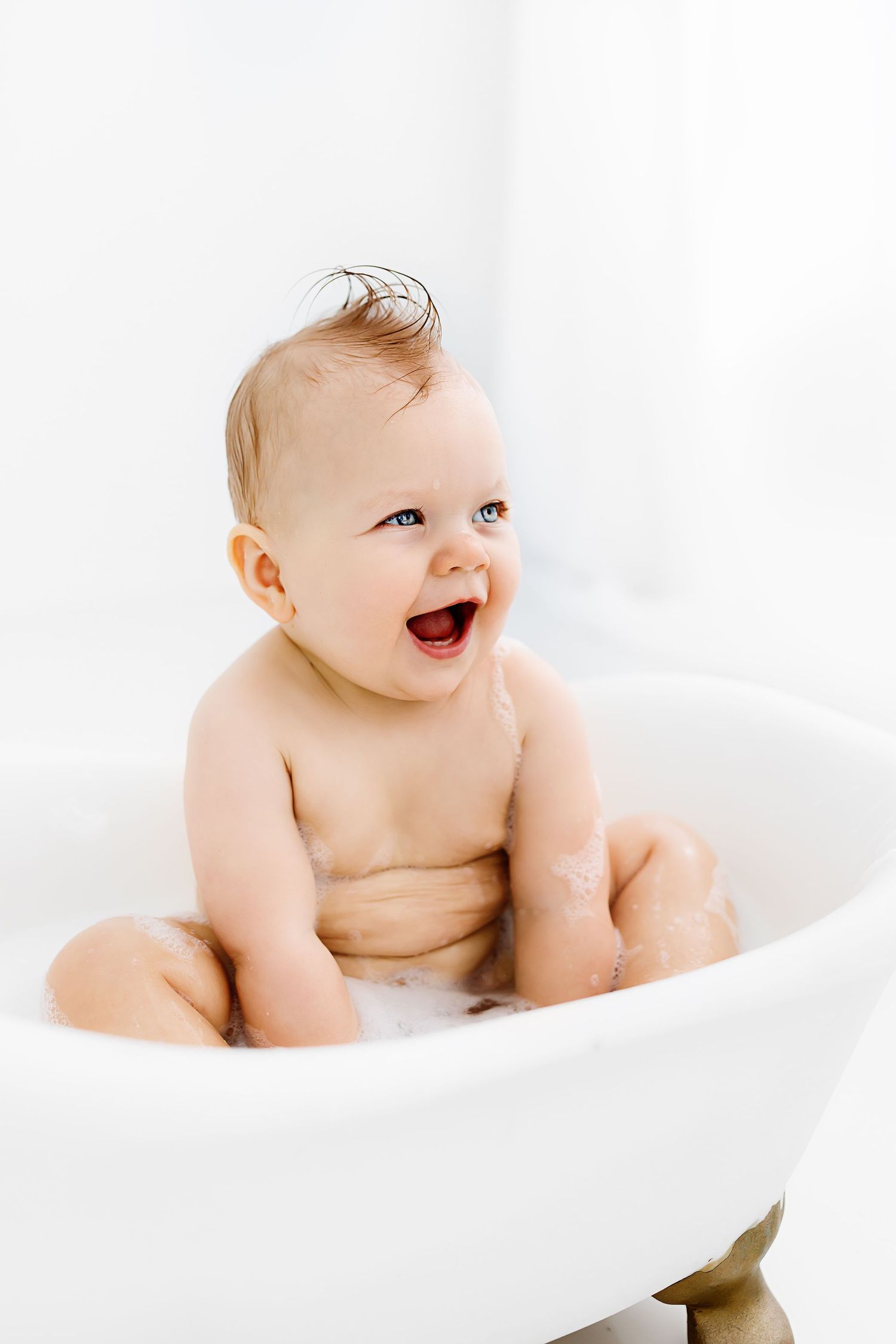 Adorable baby laughing while sitting in white bathtub with bright background.