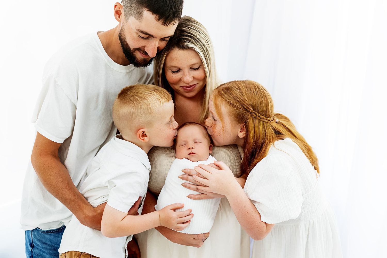 A warm family moment as parents and siblings gather close together, sharing affectionate smiles with a baby in white.