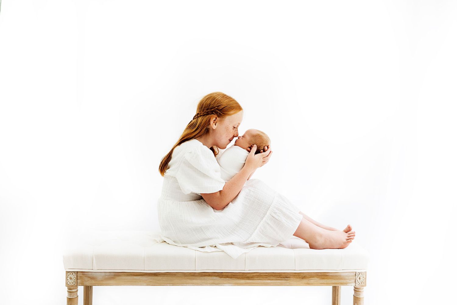 A parent in white clothing sits on a bright bench tenderly holding their newborn baby against a white background.