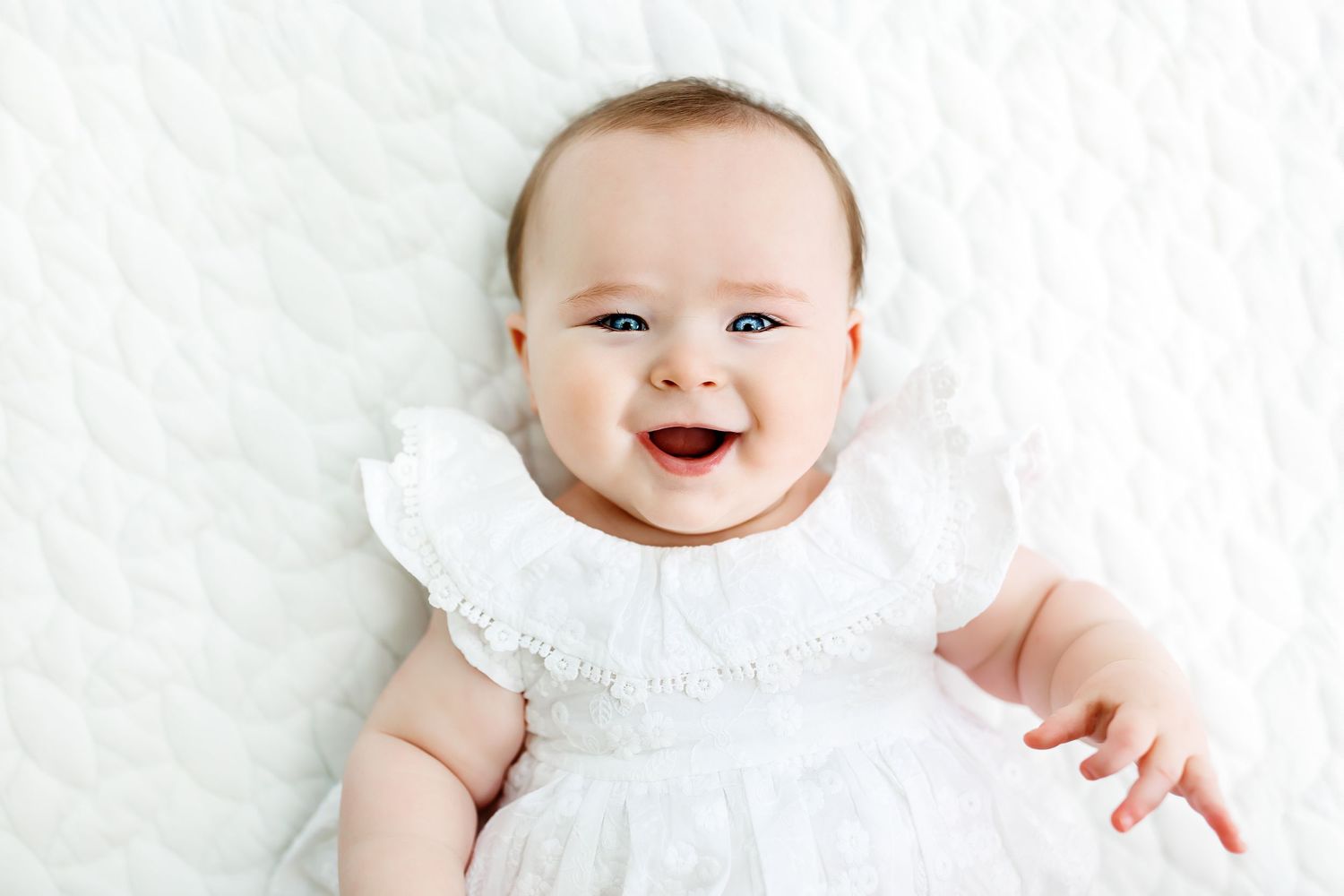 A happy baby in a white outfit lies on a textured white blanket while smiling at the camera.